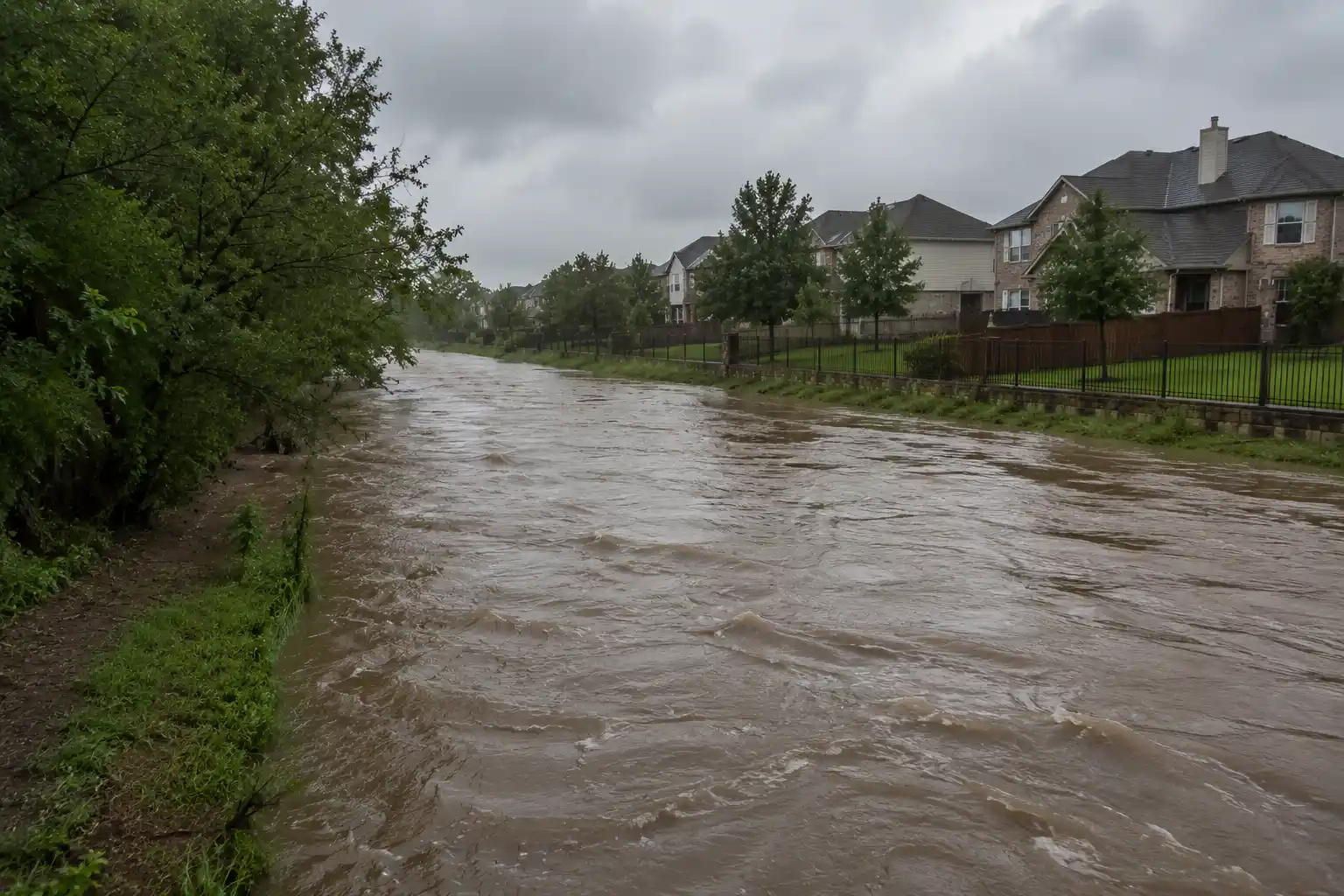 Creek flooding near Frisco homes after heavy rain, with elevated muddy water levels and an overcast sky.