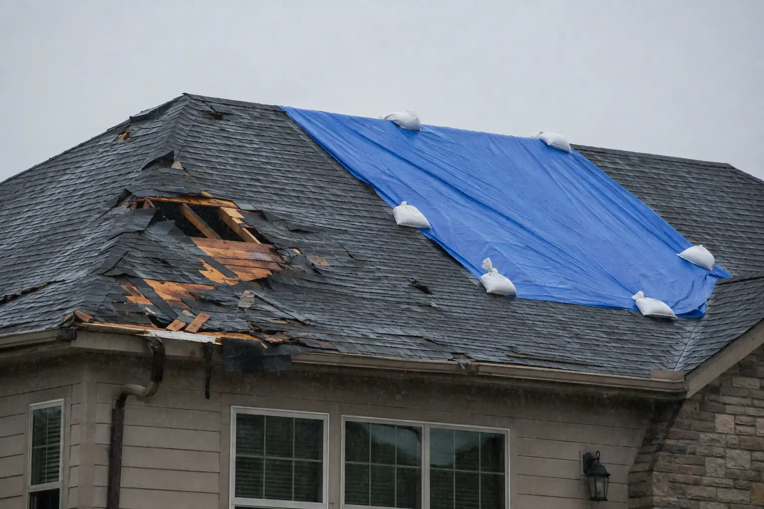 Residential rooftop with storm damage and a blue tarp covering part of the roof during rainy, overcast weather.